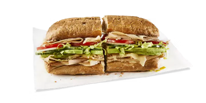 A fresh Potbelly Menu item, the Avo Turkey, featuring a toasted sub roll filled with hand-sliced turkey breast, melted Swiss cheese, fresh-sliced avocado, and crisp cucumbers, layered with lettuce and tomatoes, cut into portions and served on white paper against a black background, priced at $12.29 with 642 calories.