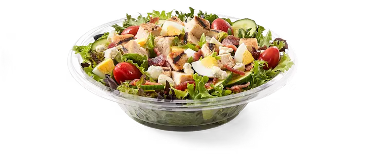 A vibrant Potbelly Menu item, the Potbelly Farmhouse Salad, featuring a generous bowl of field greens topped with all-natural grilled chicken breast, crispy bacon, crumbled blue cheese, hard-boiled egg, fresh cucumber, and juicy tomatoes, drizzled with creamy buttermilk ranch dressing, served in a clear bowl against a black background, priced at $10.99 with 425 calories.
